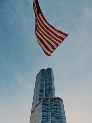 A tall skyscraper rises into the sky, made of glass with a sleek modern design. An American flag waves prominently above, contrasting with the blue sky.