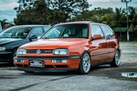 A bright orange Volkswagen GTI is parked on a paved surface alongside a black car. The car is in a serene outdoor environment with trees and cloudy skies in the background. The GTI has distinctive alloy wheels and a front license plate that reads 'VipCar Bergamini Estética Automotiva'.