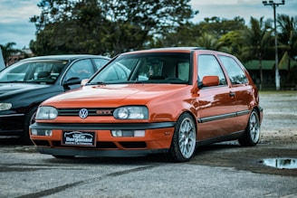 A bright orange Volkswagen GTI is parked on a paved surface alongside a black car. The car is in a serene outdoor environment with trees and cloudy skies in the background. The GTI has distinctive alloy wheels and a front license plate that reads 'VipCar Bergamini Estética Automotiva'.