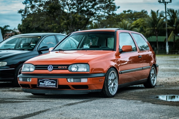 A bright orange Volkswagen GTI is parked on a paved surface alongside a black car. The car is in a serene outdoor environment with trees and cloudy skies in the background. The GTI has distinctive alloy wheels and a front license plate that reads 'VipCar Bergamini Estética Automotiva'.