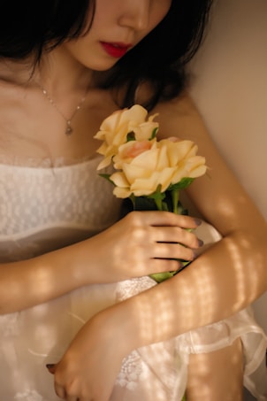 A person in a white dress holds a bouquet of light yellow roses. Soft lighting creates a patterned shadow effect over the scene, conveying a serene and intimate atmosphere. The image composition focuses on the delicate features of the dress and flowers, with the person's face partially visible and lips accentuated with red lipstick.
