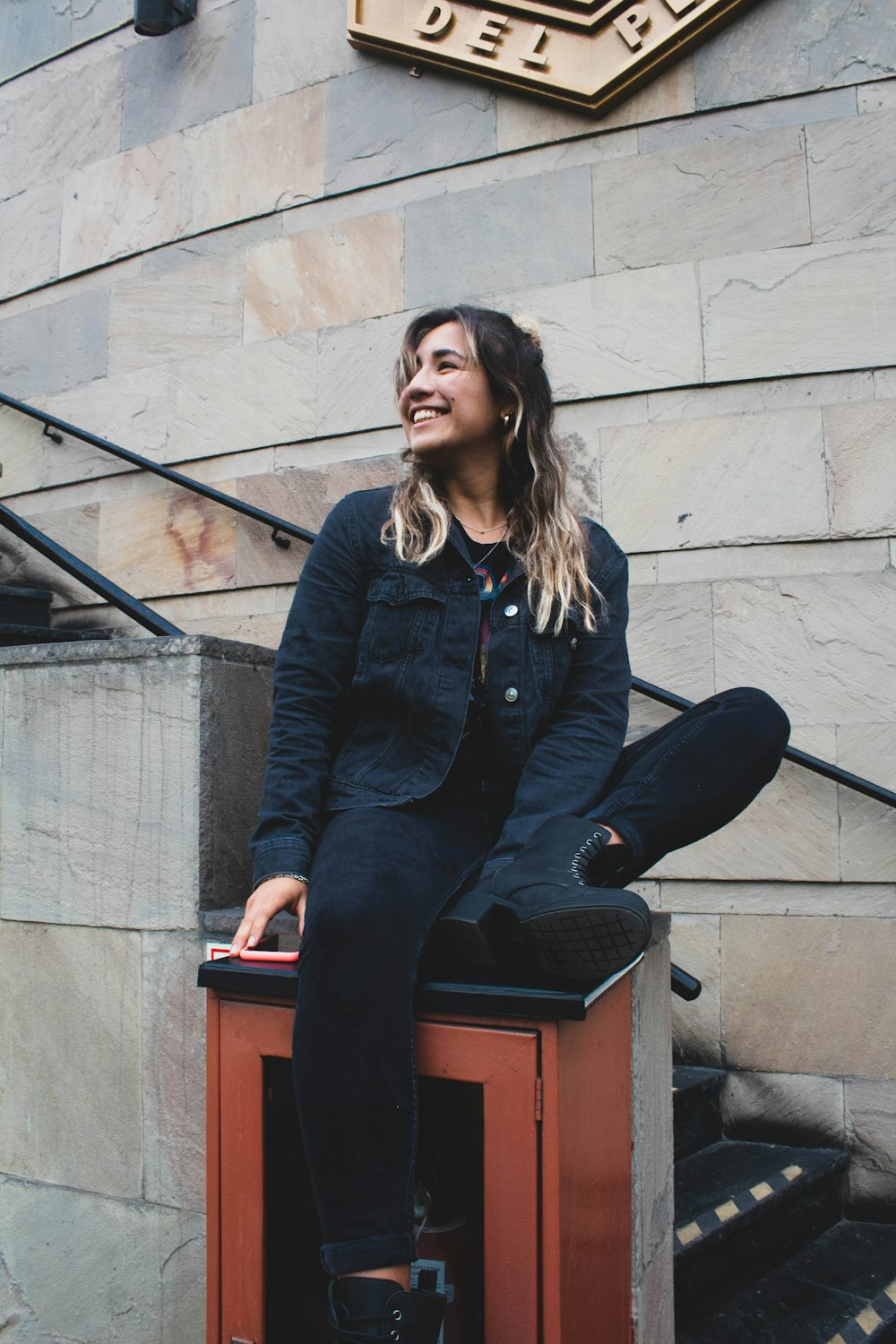 smiling woman in blue jacket and black denim jeans sitting on top of brown wall mounted box