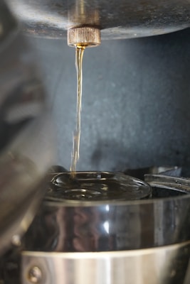 A close-up view of liquid pouring from a metallic spout into a transparent container below, possibly a glass pot. The liquid appears to be a clear, slightly amber color. The image hints at the process of brewing or distillation, with a focus on the fluid's flow and collection.