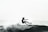 A surfer expertly balances on a surfboard amidst crashing waves, with water splashing around. The scene conveys a sense of motion and skill against the backdrop of the ocean.