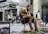A street musician sits on a stool playing a guitar-like instrument with a microphone in front of him. He is wearing a brown fur-lined jacket and boots. Several pedestrians walk by in the background, including a woman holding a tote bag and a man wearing a hat. Posters and equipment are visible near the musician.