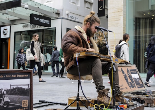 A street musician sits on a stool playing a guitar-like instrument with a microphone in front of him. He is wearing a brown fur-lined jacket and boots. Several pedestrians walk by in the background, including a woman holding a tote bag and a man wearing a hat. Posters and equipment are visible near the musician.
