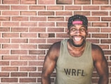 A person with a muscular build is wearing a tank top labeled 'WRFL' and a backward baseball cap. They are standing in front of a brick wall, smiling broadly and exuding a joyful expression.