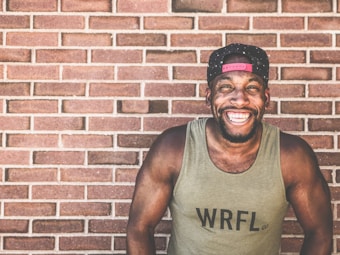 A person with a muscular build is wearing a tank top labeled 'WRFL' and a backward baseball cap. They are standing in front of a brick wall, smiling broadly and exuding a joyful expression.