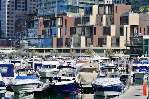 A marina filled with various docked boats and yachts sits in front of a large, modern building complex. The architecture features a mix of glass and brown concrete with balconies overlooking the waterfront. The area is bustling with activity as people walk around the boardwalks among palm trees lining the marina.