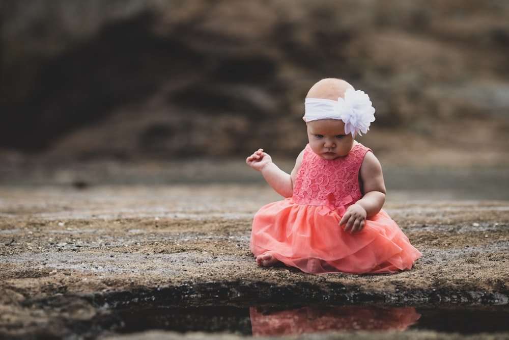 shallow focus photo of toddler in pink sleeveless dress sitting near body of water