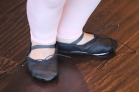 A pair of small feet in black ballet shoes stand on a wooden floor. The legs are covered in light pink leggings, suggesting they belong to a child.