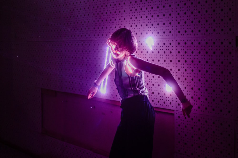 woman in grey tank top and black skirt leaning on wall with lighted pink bulbs