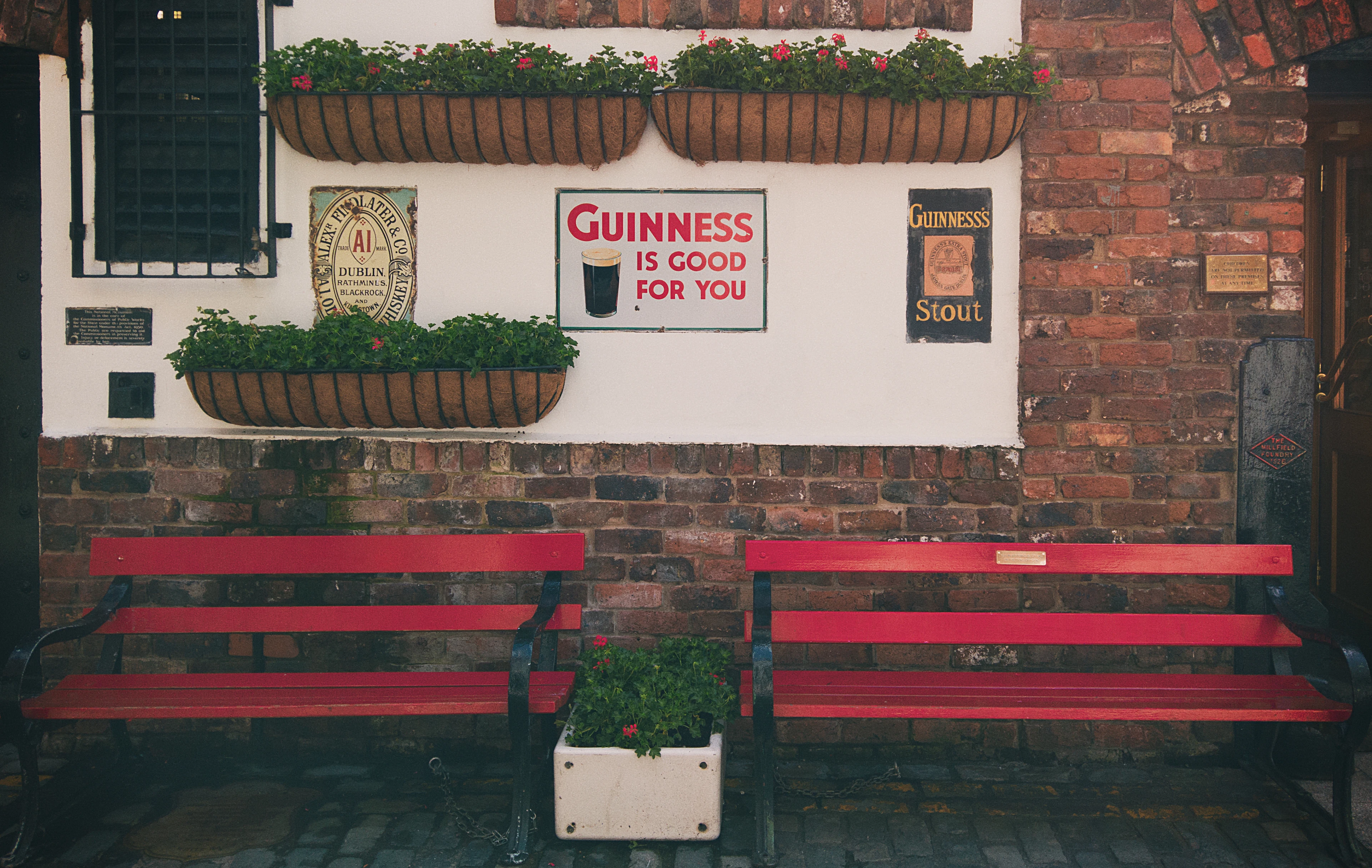 Red benches and hanging flower baskets outside a whitewashed Belfast pub decorated with vintage Guinness signs.