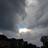 Dark storm clouds fill the sky, with a small patch of blue and white sky visible in the middle. Below, the silhouette of buildings and trees can be seen, along with power lines running across the scene.