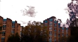 A set of multi-story residential buildings is surrounded by trees. The buildings have an angular architecture with a facade of tan bricks and blue trim. The sky is overcast with a few scattered clouds.