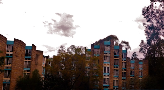 A set of multi-story residential buildings is surrounded by trees. The buildings have an angular architecture with a facade of tan bricks and blue trim. The sky is overcast with a few scattered clouds.