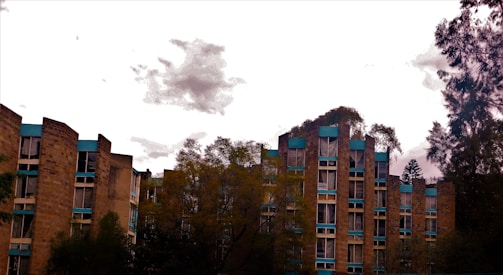 A set of multi-story residential buildings is surrounded by trees. The buildings have an angular architecture with a facade of tan bricks and blue trim. The sky is overcast with a few scattered clouds.