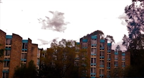 A set of multi-story residential buildings is surrounded by trees. The buildings have an angular architecture with a facade of tan bricks and blue trim. The sky is overcast with a few scattered clouds.