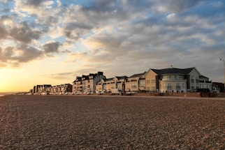 A row of seaside buildings with a classic architectural style lines a pebble beach. The sky is partially cloudy, illuminated by a warm sunset, casting a golden hue over the scene.
