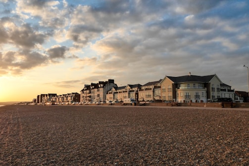 A row of seaside buildings with a classic architectural style lines a pebble beach. The sky is partially cloudy, illuminated by a warm sunset, casting a golden hue over the scene.