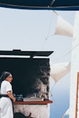 An elderly person dressed in traditional attire stands next to an outdoor oven. Freshly baked bread is visible on a tray. White walls and sails, along with a glimpse of the ocean in the background, suggest a coastal setting.