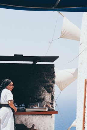 An elderly person dressed in traditional attire stands next to an outdoor oven. Freshly baked bread is visible on a tray. White walls and sails, along with a glimpse of the ocean in the background, suggest a coastal setting.