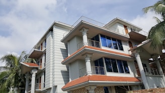 A modern multi-story house with intricate architectural details, featuring white walls and columns. The building has multiple balconies with metal railings and large blue-tinted windows. The roof is made of red tiles, and several palm trees surround the structure, adding a tropical feel. The sky is partly cloudy.
