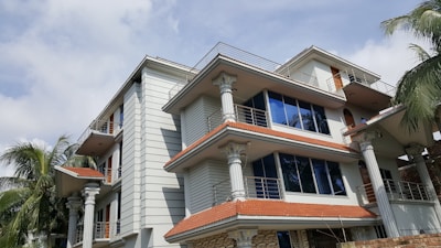 A modern multi-story house with intricate architectural details, featuring white walls and columns. The building has multiple balconies with metal railings and large blue-tinted windows. The roof is made of red tiles, and several palm trees surround the structure, adding a tropical feel. The sky is partly cloudy.
