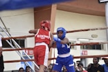 Two boxers are engaged in a match inside a boxing ring, surrounded by ropes. One boxer is wearing a red uniform and helmet, while the other is wearing blue gear. The audience in the background and signage on the walls suggest an indoor sports facility.