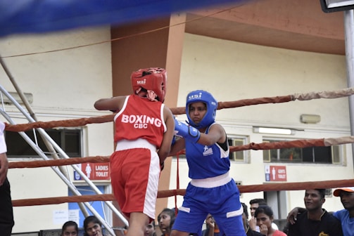 Two boxers are engaged in a match inside a boxing ring, surrounded by ropes. One boxer is wearing a red uniform and helmet, while the other is wearing blue gear. The audience in the background and signage on the walls suggest an indoor sports facility.