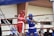 Two boxers are engaged in a match inside a boxing ring, surrounded by ropes. One boxer is wearing a red uniform and helmet, while the other is wearing blue gear. The audience in the background and signage on the walls suggest an indoor sports facility.