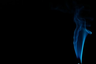 A wispy trail of blue smoke elegantly rises against a dark background, creating a contrast between the vivid color of the smoke and the deep black surroundings. The smoke appears to gracefully twist and swirl, forming gentle curves and delicate patterns.