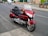 A red touring motorcycle is parked on the side of a suburban street. The motorcycle features a large windshield, comfortable seating, and storage compartments. The street is lined with residential houses, and a few cars are parked along the road.