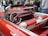 A classic convertible car interior with a vibrant red and white leather upholstery. The steering wheel and dashboard feature a vintage design, perfectly complementing the retro aesthetic of the car. In the background, a group of people stands on a street, partially visible and slightly out of focus.