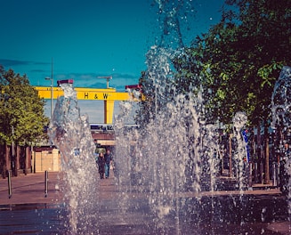 A lively urban scene with water fountains spraying upwards in the foreground. Behind the water display, there is a large yellow crane with the letters 'H & W' prominently displayed. Trees line the both sides, adding greenery, and several people are visible in the background, walking through the area.