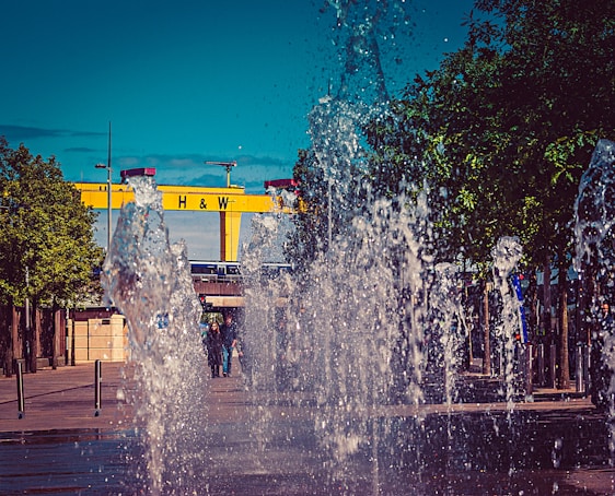A lively urban scene with water fountains spraying upwards in the foreground. Behind the water display, there is a large yellow crane with the letters 'H & W' prominently displayed. Trees line the both sides, adding greenery, and several people are visible in the background, walking through the area.