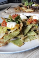 A plate of loaded nachos featuring vibrant green and yellow tortilla chips, topped with sour cream, guacamole, diced tomatoes, and olives. In the background, there are additional dishes, including what appears to be roasted root vegetables or fries.