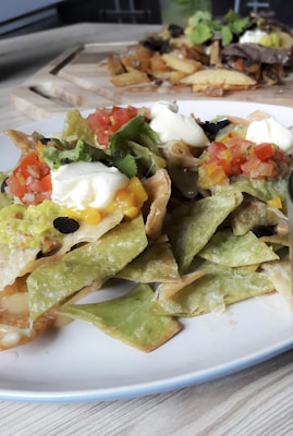 A plate of loaded nachos featuring vibrant green and yellow tortilla chips, topped with sour cream, guacamole, diced tomatoes, and olives. In the background, there are additional dishes, including what appears to be roasted root vegetables or fries.