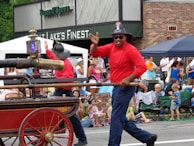 A festive parade scene includes a firefighter in a red uniform and helmet, smiling and waving to the crowd while holding onto a vintage fire vehicle with a large, ornate water hose. The background shows a diverse group of onlookers, including families and children, enjoying the event. Some are seated on portable chairs, and there is a storefront in the backdrop.