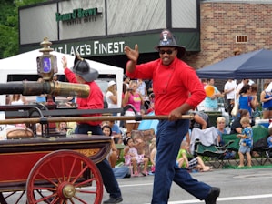 A festive parade scene includes a firefighter in a red uniform and helmet, smiling and waving to the crowd while holding onto a vintage fire vehicle with a large, ornate water hose. The background shows a diverse group of onlookers, including families and children, enjoying the event. Some are seated on portable chairs, and there is a storefront in the backdrop.