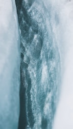 A close-up view of a crevasse within a glacier, showcasing layers of compacted ice and snow. The ice appears textured and variegated, with light filtering through, creating a bluish hue.