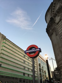 A London Underground sign stands prominently in an urban setting, surrounded by buildings with distinct architectural features. The sky is clear with a contrail visible, adding a dynamic element to the scene. Street lights and signage are present, indicating an active, city atmosphere.