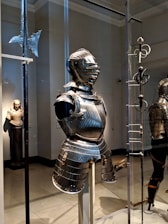 A suit of medieval armor is displayed in a museum setting, surrounded by other historical artifacts such as a polearm weapon and a rapier. The armor is intricately designed with ridges and metallic surfaces that reflect the lighting of the room. In the background, there is a statue of a robed figure, adding to the historical ambiance.