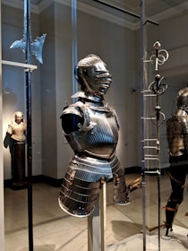 A suit of medieval armor is displayed in a museum setting, surrounded by other historical artifacts such as a polearm weapon and a rapier. The armor is intricately designed with ridges and metallic surfaces that reflect the lighting of the room. In the background, there is a statue of a robed figure, adding to the historical ambiance.