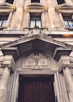 A grand neoclassical building entrance with ornate columns and detailed architectural features. The facade includes intricately carved stonework, emphasizing classical elements. Prominent numerals 'No 16' and the caduceus symbol are displayed above large wooden doors.