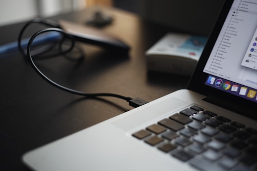 A close-up of a laptop with an open application visible on the screen. A USB cable is plugged into the laptop, with a blurred object that appears to be an external hard drive in the background. The setting is a dimly-lit environment, suggesting a workspace.