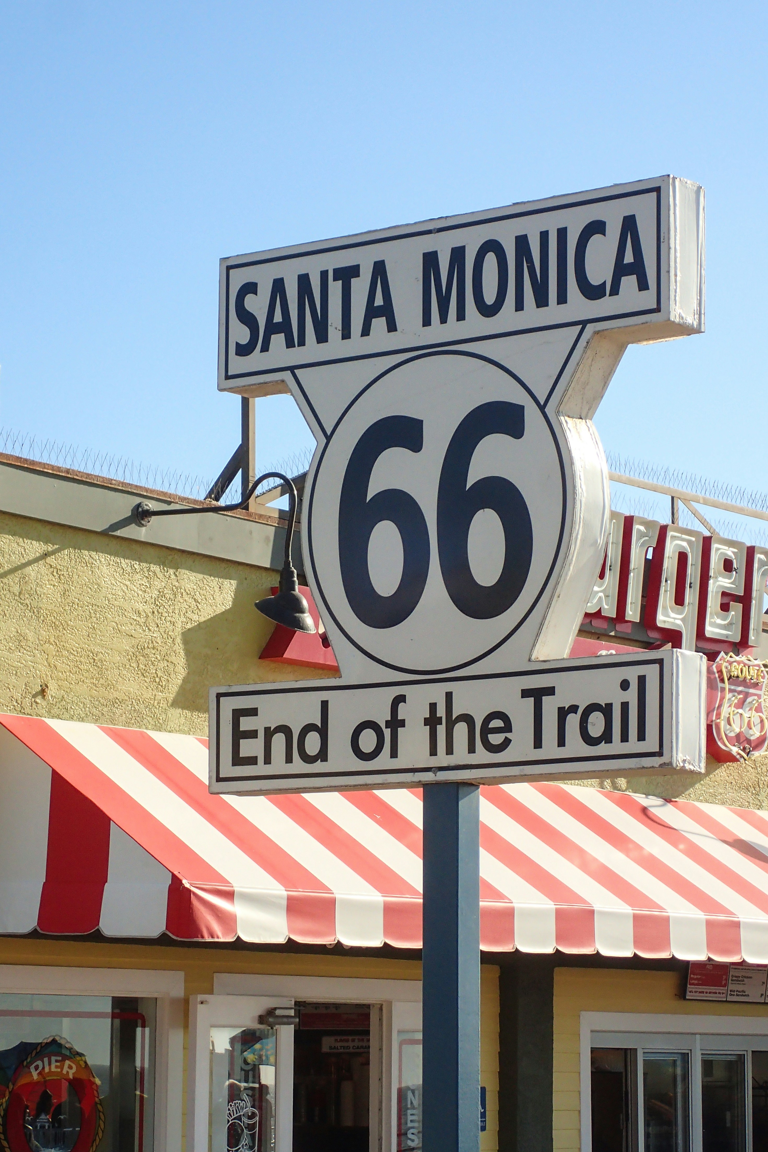 Route 66 sign reading End of the Trail rises above a sunlit Santa Monica storefront with a red-and-white striped awning.