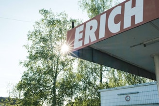 A large sign with the partial word 'ERICH' is mounted on a corner roof, slightly weathered. The sun shines through the leafy branches of tall trees in the background, casting natural light. Below the sign, part of a building with tiled walls and a traffic height restriction sign is visible.