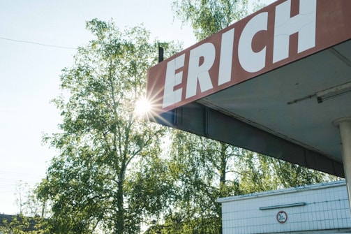 A large sign with the partial word 'ERICH' is mounted on a corner roof, slightly weathered. The sun shines through the leafy branches of tall trees in the background, casting natural light. Below the sign, part of a building with tiled walls and a traffic height restriction sign is visible.