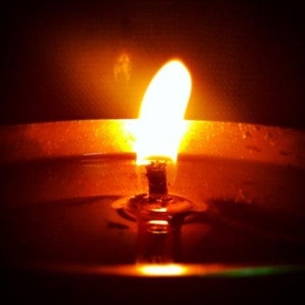 A close-up view of a lit candle with a bright flame surrounded by a soft glow, casting a warm and inviting ambiance. The wick is clearly visible, and there is a pool of melted wax around it.
